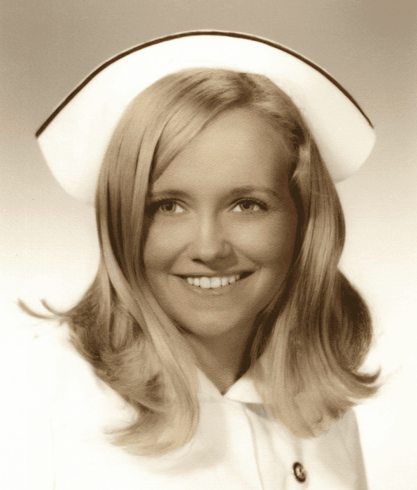 A young woman with long blonde hair wears a nurse's uniform and cap, smiling at the camera in this vintage, sepia-toned portrait.