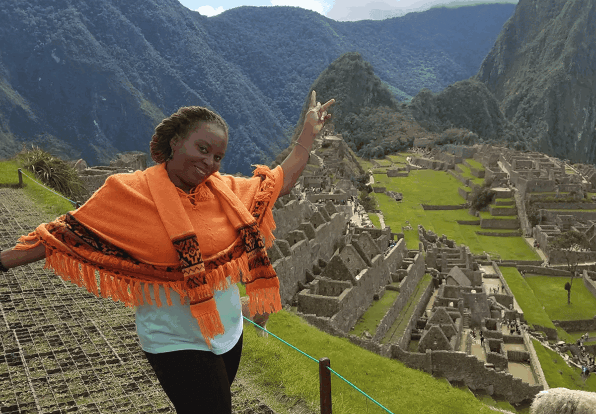 A woman in an orange poncho poses and smiles with arms raised at Machu Picchu, with stone ruins and mountains visible in the background.