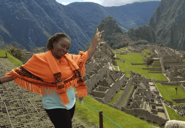 A woman in an orange poncho poses and smiles with arms raised at Machu Picchu, with stone ruins and mountains visible in the background.