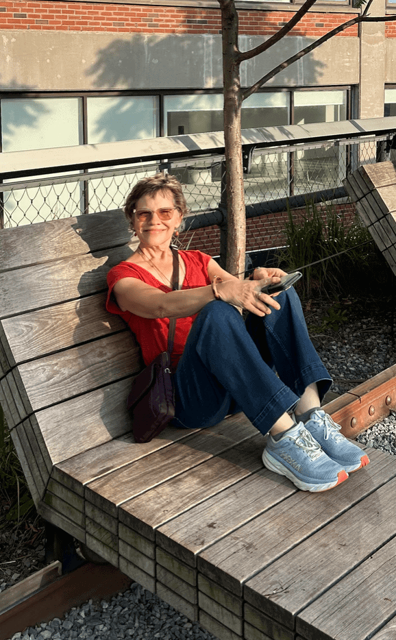 A woman wearing sunglasses, a red shirt, and blue jeans sits on a wooden bench outdoors, holding a smartphone with one hand.