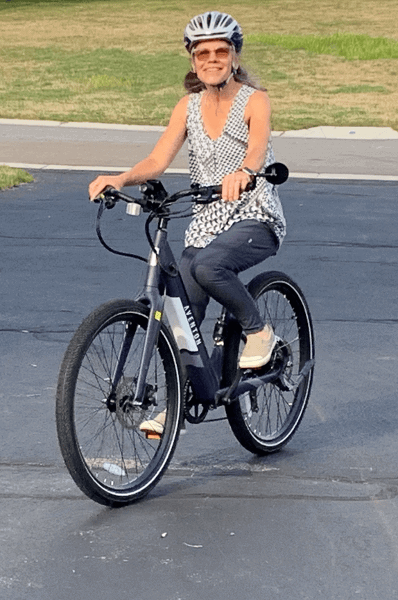 A woman wearing a helmet, sunglasses, and sleeveless top rides a bicycle on a paved surface with grass in the background.