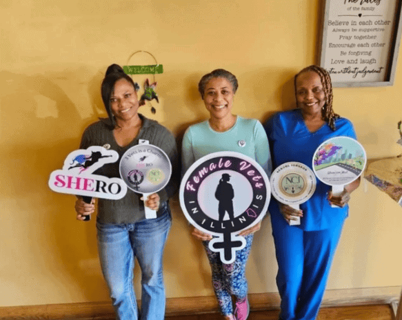 Three women stand indoors holding signs for organizations including "SHERO" and "Female Vets in Illinois," smiling for the camera.