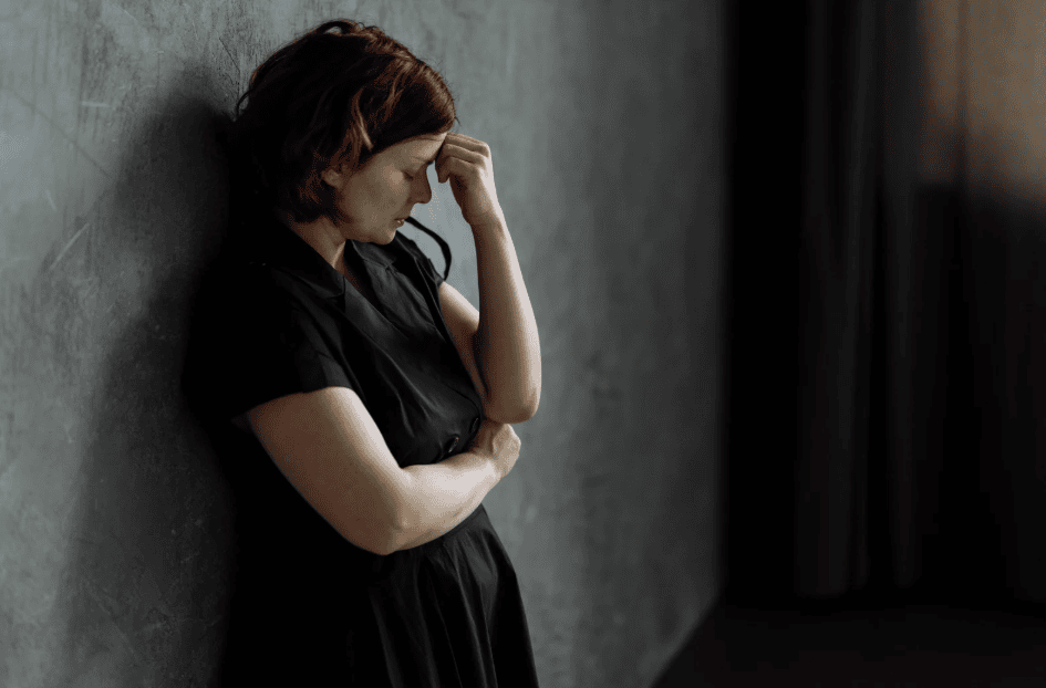 A woman in a black dress stands against a gray wall with her head resting on her hand, appearing deep in thought or concerned.