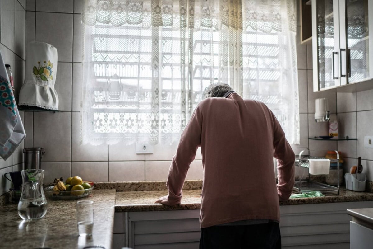 An older person stands leaning on a kitchen counter, facing a window with lace curtains. Fruit, dishes, and a pitcher are visible on the counter.