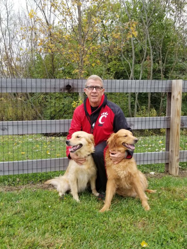 An older man in a red and black jacket kneels on grass, smiling with two golden retrievers in front of a wooden fence and trees.