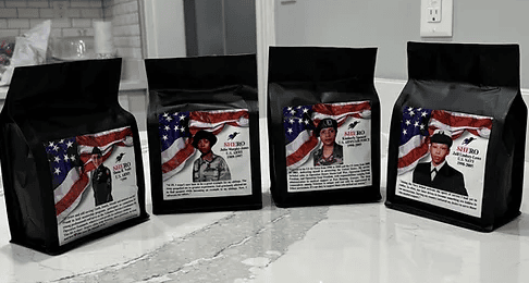 Four black coffee bags sit on a countertop, each featuring a photo of a different person in military uniform with an American flag backdrop on the label.