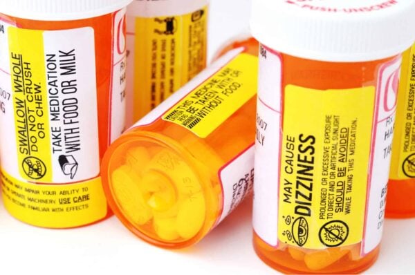 Close-up of several orange prescription pill bottles with yellow warning labels, including instructions to take with food or milk and caution about possible dizziness.