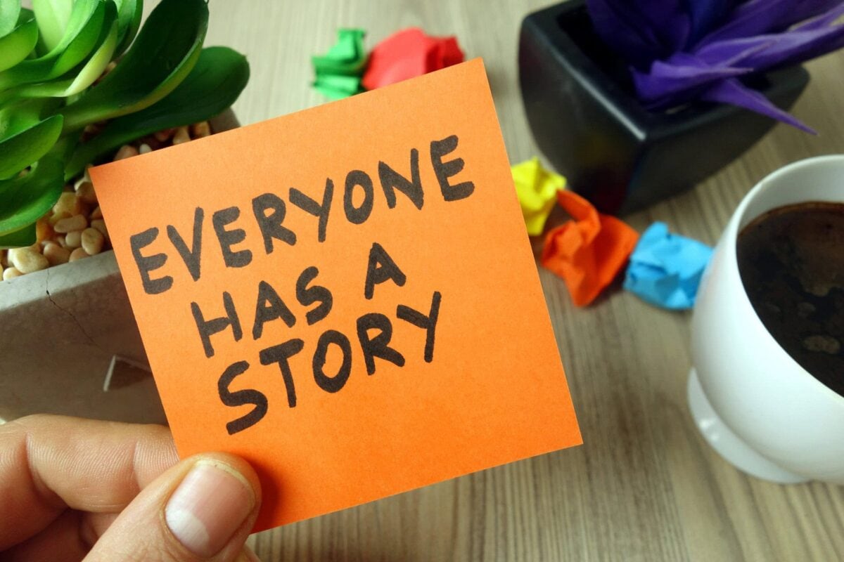 A hand holds an orange sticky note with the words "EVERYONE HAS A STORY" written in black marker, next to a coffee cup, plant, and colorful crumpled papers.