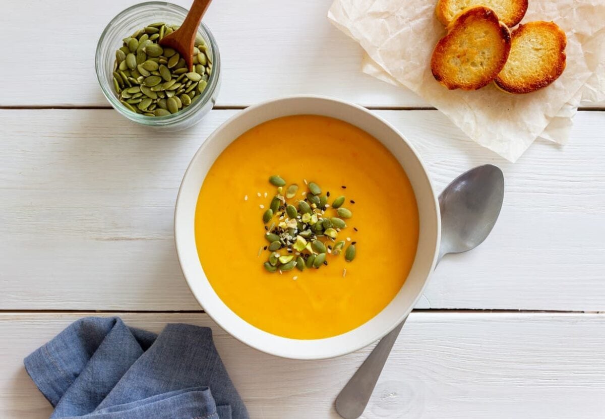 A bowl of orange-colored soup topped with seeds, a spoon beside it, a jar of pumpkin seeds with a wooden spoon, and toasted bread on parchment paper.