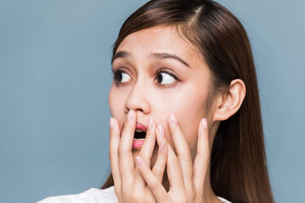 A young woman with long brown hair looks to the side with a surprised expression, covering her mouth with her hands against a plain blue background.