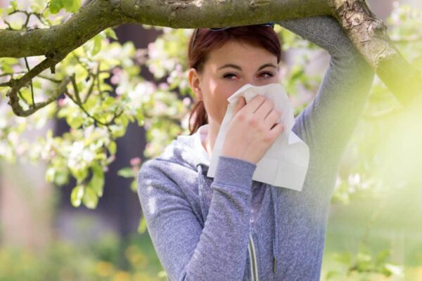 A woman standing outdoors under a tree uses a tissue to wipe or blow her nose, possibly due to allergies.