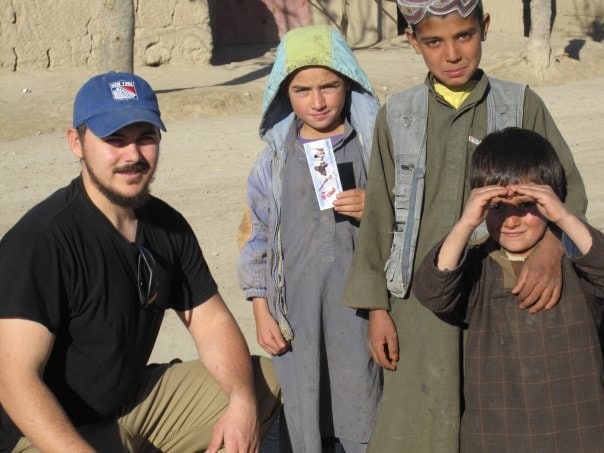 An adult man kneels beside three local children in traditional clothing outdoors; one child holds a card, and another forms binoculars with his hands.