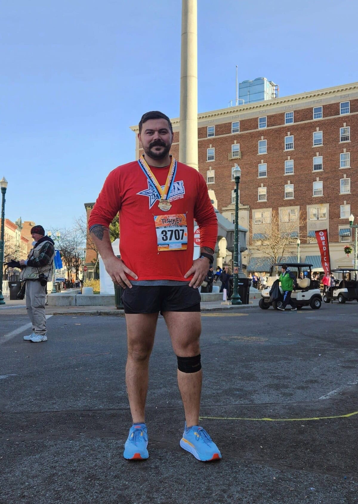 A runner wearing a red shirt, race bib 3707, and medals stands in the street after a race, with buildings and other people in the background.