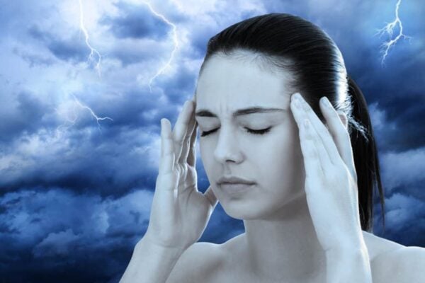 A woman with closed eyes and a pained expression holds her temples, with dark storm clouds and lightning in the background.