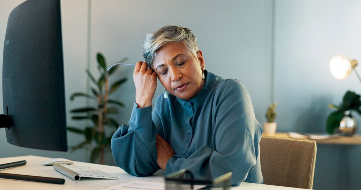 A woman sits at a desk with her eyes closed and head resting on her hand, appearing tired while working at a computer.