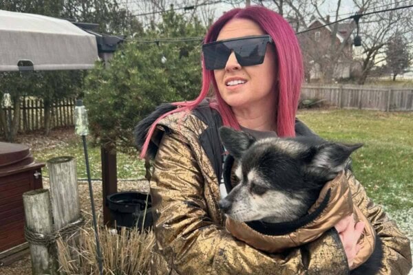 A woman with long pink hair and large sunglasses holds a black and white dog while standing outside in a backyard.