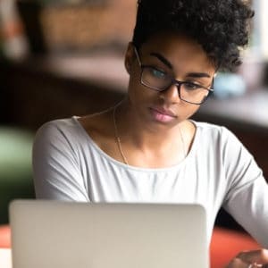A person wearing glasses and a light gray shirt is sitting indoors, looking at a laptop screen with a focused expression.