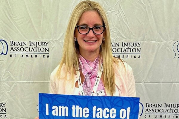 A woman wearing glasses and a pink scarf smiles while holding a blue sign in front of a Brain Injury Association of America backdrop.