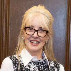 A woman with long blonde hair, glasses, and a black-and-white patterned top over a white collared shirt, smiling in front of a wooden background.