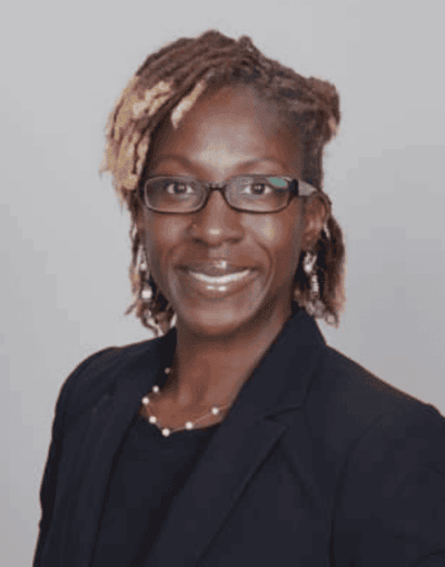 A woman wearing glasses, a dark blazer, and a necklace, smiling in a professional headshot against a plain light background.