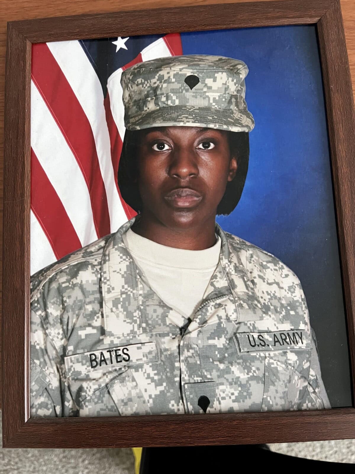 A framed photo of a U.S. Army soldier in camouflage uniform and cap, with a name tag reading "BATES," posing in front of an American flag and blue background.