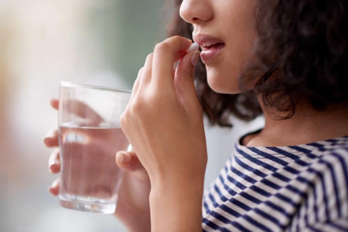 A person holding a glass of water brings a pill to their mouth, preparing to take medication.