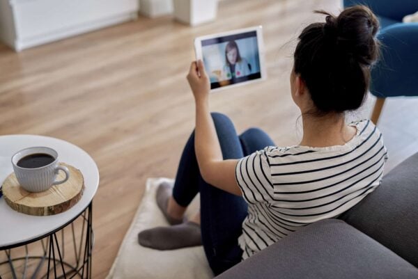 A person sitting on the floor by a couch, holding a tablet and video calling another person, with a cup of coffee on a small table nearby.