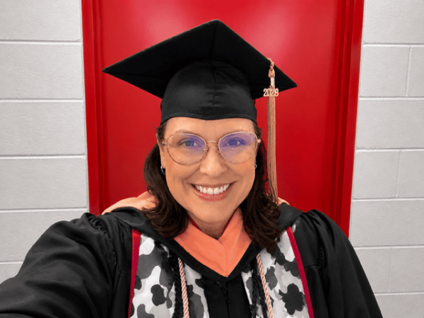 A woman with glasses, smiling with a graduation cap and gown. A red door is visible in the background