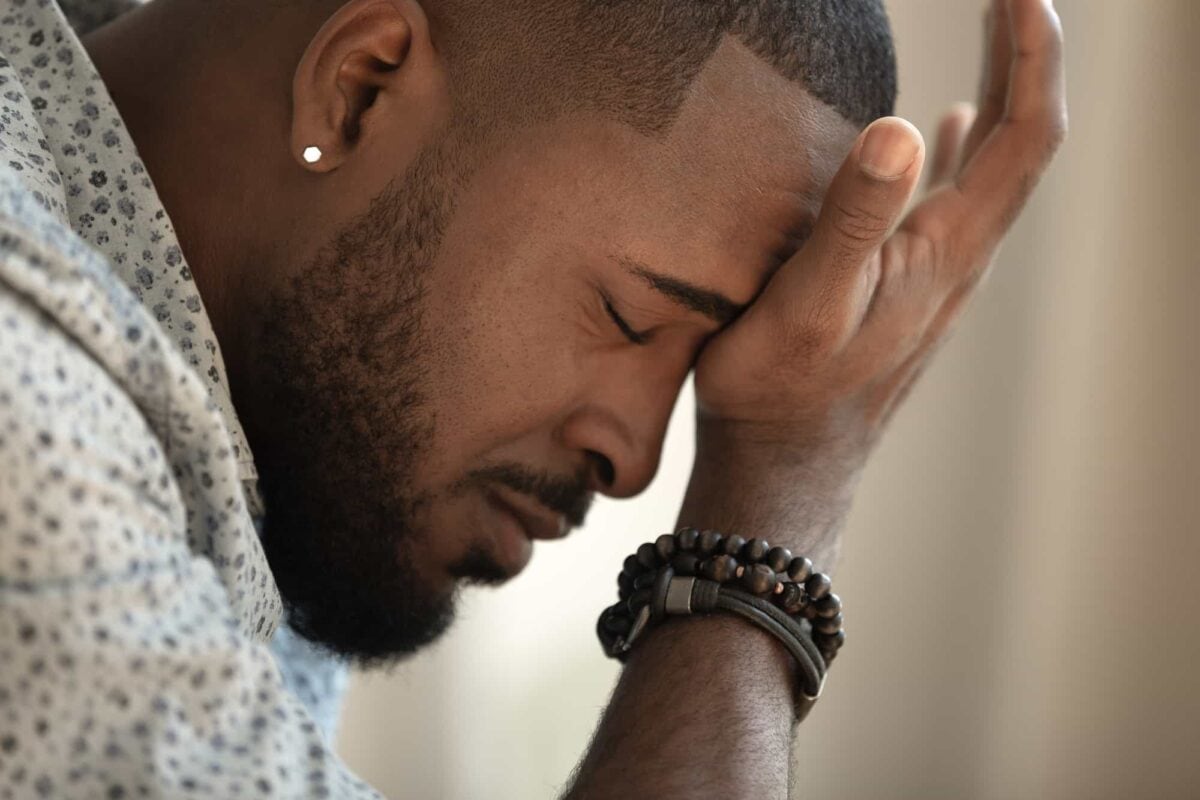 A man with closed eyes rests his forehead on his hand, appearing stressed or upset, wearing a patterned shirt, bracelets, and an earring.