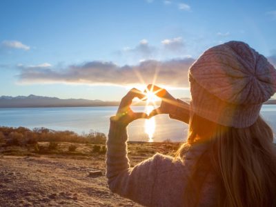 A person in a knit hat forms a heart shape with their hands, framing the sun over a lake at sunrise or sunset.