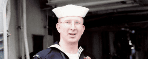 A person in a U.S. Navy uniform and white sailor cap stands indoors, facing the camera.