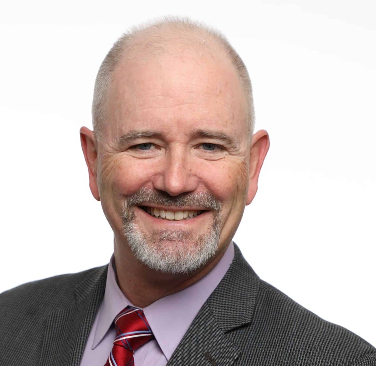 Bald man with a gray beard wearing a gray suit, lavender shirt, and red striped tie, smiling at the camera against a plain white background.