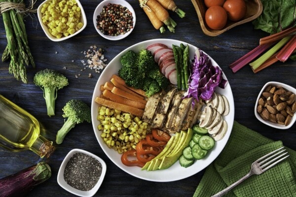 A white plate with grilled tofu, broccoli, carrots, corn, radish, cabbage, mushrooms, cucumber, tomato, bell pepper, and avocado, surrounded by various vegetables and ingredients.