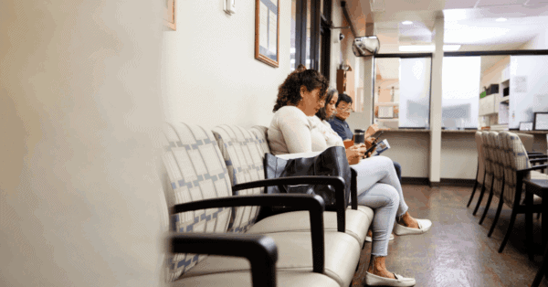 Three people sit in a row on chairs in a waiting room, looking at their phones, with an empty reception area visible in the background.