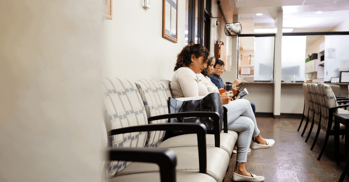 Three people sit in a row on chairs in a waiting room, looking at their phones, with an empty reception area visible in the background.