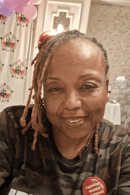 A woman with dreadlocks smiles at the camera, wearing a camouflage shirt and a red button that reads "I SUPPORT WORKERS." Balloons and a patterned backdrop are visible behind her.