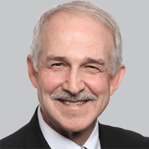 Older man with gray hair, mustache, and a slight smile, wearing a dark suit and white shirt, posing against a plain gray background.