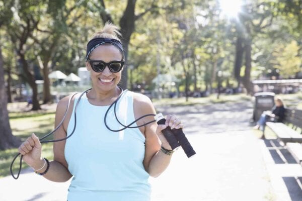 Woman wearing sunglasses and workout clothes smiles while holding a jump rope in a sunny park with trees and benches in the background.