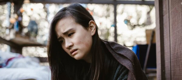 A young person with long dark hair looks downward with a concerned expression, sitting indoors near a window with blurred outdoor light in the background.