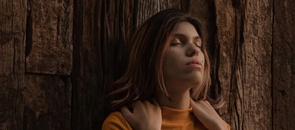 A woman with long brown hair, wearing an orange top, stands against a wooden wall with her eyes closed and hands near her neck.