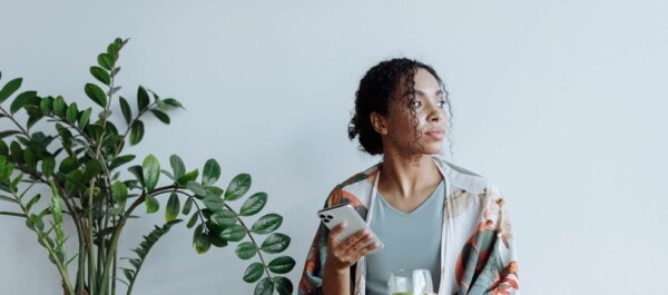 A woman holding a smartphone sits indoors next to a large green plant, looking off to the side.