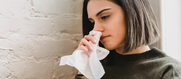 Woman with short dark hair holds a tissue to her nose, appearing to be unwell, standing indoors near a white brick wall.