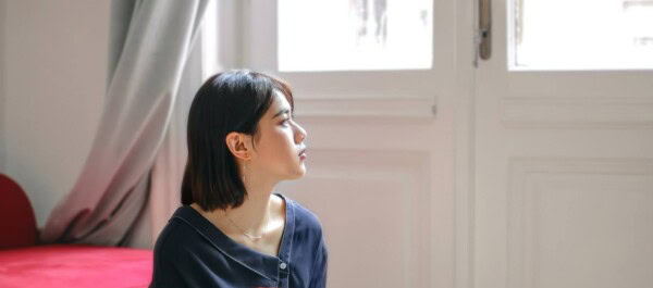 A woman with short dark hair sits indoors, looking to the side near a window with white curtains and a white door in the background.