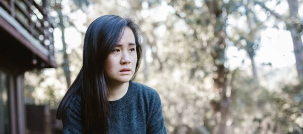 A woman with long dark hair and a gray sweater sits outdoors, looking thoughtful and slightly concerned, with blurred trees in the background.
