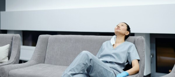 A person in medical scrubs sits on a gray couch, leaning back and looking up, with a mask hanging from one ear.