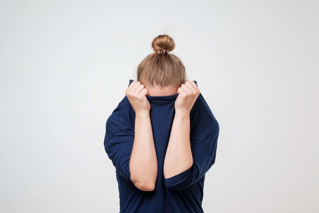 Person with hair in a bun pulls a navy shirt over their face, standing against a plain light background.