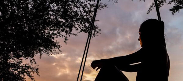A person sits on a swing at sunset, silhouetted against a colorful sky with tree branches framing the scene.