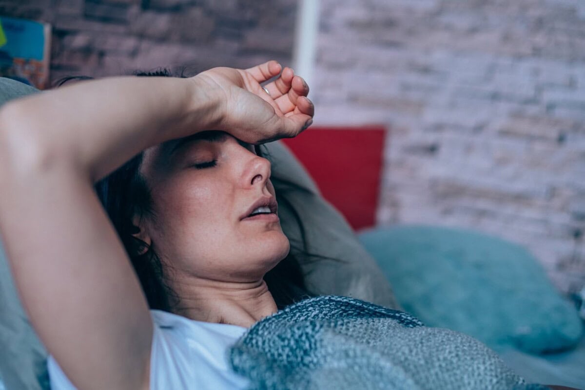 A woman lies on a couch with her eyes closed and hand on her forehead, appearing tired or unwell.