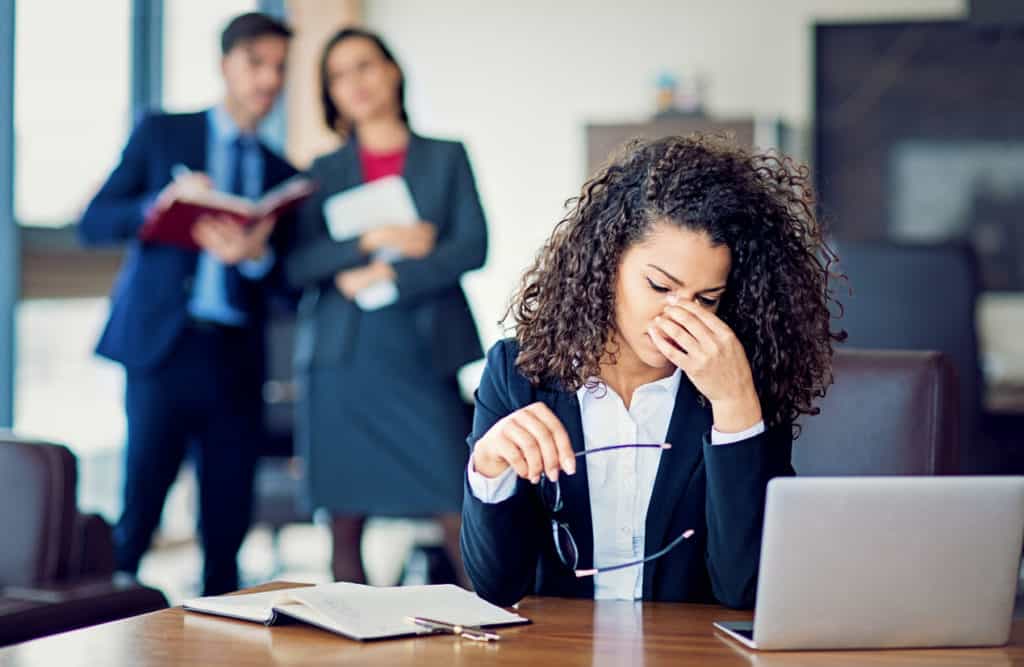 A woman in business attire sits at a desk looking stressed, holding her glasses, with two colleagues standing and talking in the background.
