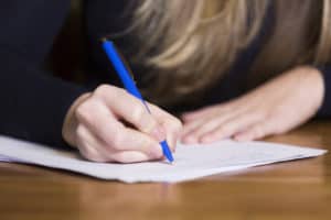 A person with long hair writes on paper with a blue pen at a wooden table.